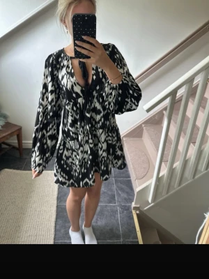 Svartvit kortklänning med mönster - Säljer en svartvit kortklänning med grafiskt mönster och långa ärmar. Klänningen har v-ringning med snörning och en lös passform som ger en avslappnad look. Perfekt för dig som gillar statement-plagg och vill sticka ut.