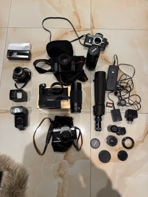 Nikon systemkamerapaket med tillbehör - Säljer ett komplett Nikon kamerakit med flera analoga systemkameror, objektiv, blixtar, stativ, batteriladdare och olika filter. Allt är svart och silverfärgat, mestadels i metall och plast. Perfekt för dig som vill börja med analog foto eller vill ha extra utrustning till din samling.