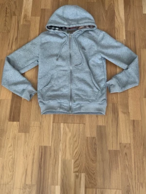 Grå hoodie med dragkedja Burberry - Snygg grå hoodie från Burberry med dragkedja framtill och klassiskt rutigt foder på insidan. Hoodien har två fickor fram, ribbade muddar och justerbar huva med snören. Perfekt för en chill och stilren look.