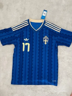 Blå Sverige tröja från Adidas - Säljer en blå fotbollströja från Adidas med Sveriges landslagsemblem och nummer 17 i gult. Tröjan har beige detaljer, klassiska Adidas-ränder på axlarna och ett unikt mönster över hela tyget. Perfekt för dig som älskar fotboll och vill stötta Sverige!