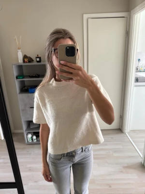 Vit kortärmad stickad tröja - Säljer en enkel och stilren vit stickad tröja med korta ärmar och rund hals. Tröjan har en boxig, croppad passform och ribbade kanter vid ärmslut och nederkant. 
