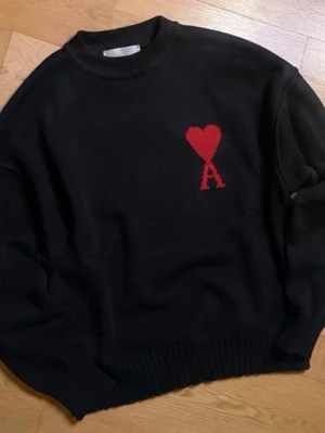 Svart stickad tröja Ami Paris - Svart stickad tröja från Ami Paris med röd hjärtlogga och bokstaven A på bröstet. Tröjan har rund halsringning, ribbade muddar och avslut. Perfekt oversized passform för en avslappnad look.