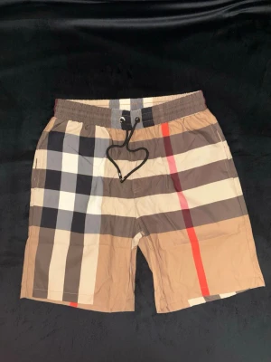 Burberry shorts - Snygga rutiga shorts från Burberry i beige, svart, vit och röd. Klassisk Burberry-ruta med elastisk midja och snörning framtill. Perfekta för sommaren och ger en trendig vibe till din outfit. Storlek M, helt nya med tags på.
