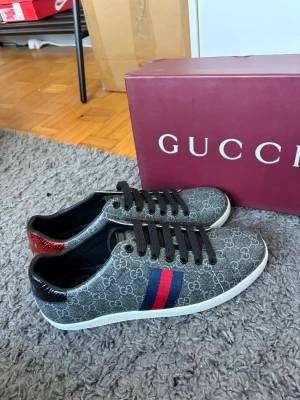 Gucci GG Supreme sneakers grå/röd - Snygga Gucci sneakers i grått GG Supreme-mönster med klassiska blå och röda ränder på sidan. Skorna har svarta skosnören, vit sula och detaljer i rött och svart på hälen. Tillverkade i canvas med läderdetaljer. Perfekta för dig som vill sticka ut med lyxig streetstyle.