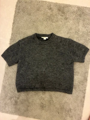 Grå stickad tröja Gina Tricot - Mysig grå stickad tröja från Gina Tricot med korta ärmar och rund halsringning. Tröjan har en enkel, rak passform och är tillverkad i ett mjukt, fluffigt material som passar perfekt för kyligare dagar.