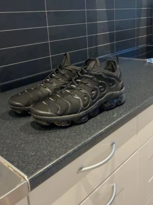 Nike Air VaporMax Plus svarta sneakers - Svarta Nike Air VaporMax Plus sneakers med en futuristisk design och synliga luftbubblor i sulan. Ovandelen är i textil och syntet med vågformade detaljer och diskret Swoosh-logga. Skorna har snörning och en chunky sula som ger en sportig vibe.