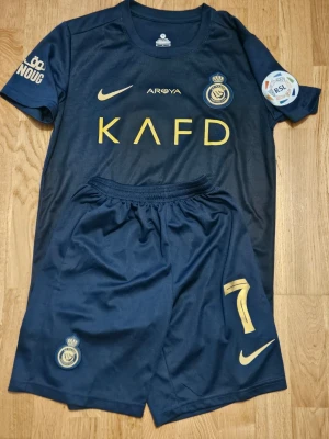 Al Nassr matchställ Nike blå/guld - Snyggt matchställ från Al Nassr FC i mörkblått med guldiga detaljer. Tröjan har klubbmärke, Nike-logga och sponsortryck framtill samt patch på ärmen. Shortsen har nummer 7 och klubbmärke. Tillverkat i lätt och funktionellt material, perfekt för fotbollsträning eller match.