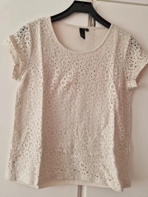 Vit spetsblus från H&M - Söt vit blus från H&M med korta ärmar och spetsdetaljer framtill och på ärmarna. Baksidan är i mjuk bomull för extra komfort. Perfekt till jeans eller kjol för en fräsch look.