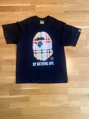 Svart BAPE t-shirt med rutigt tryck - Svart t-shirt från BAPE med det klassiska apansikte-trycket i rutigt mönster på bröstet och texten 'By Bathing Ape' under. Rund halsringning och korta ärmar. Perfekt statement-plagg för streetwear-stilen.