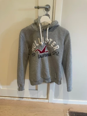 Grå hoodie från Hollister - Snygg grå hoodie från Hollister med broderad logga och texten 'California 1922' på bröstet. Hoodien har en stor huva med vita dragsnören och mjukt material på insidan. Perfekt för chill dagar och streetwear-stil.