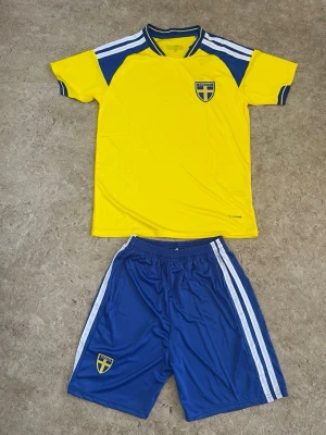 Svenskt fotbollset i gul och blå - Säljer ett fotbollset med gul t-shirt och blå shorts, båda med Sveriges landslagsemblem. Tröjan har blå detaljer och vita ränder på axlarna, medan shortsen har vita ränder längs sidorna. Materialet är lätt och andas, perfekt för träning eller match.