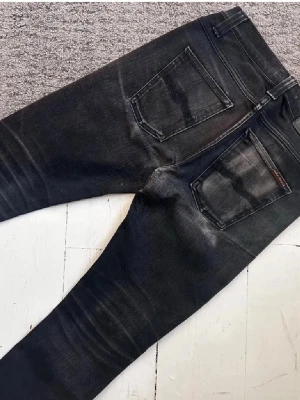 Svarta slim fit jeans från Nudie Jeans - Säljer ett par svarta slim fit jeans från Nudie Jeans med klassisk femficksmodell och snygga slitningar. Jeansen har raka ben och är tillverkade i stretchig denim för skön passform. Den är i storlek W29 och L32. Det är bara att höra av sig vid minsta fundering🙌