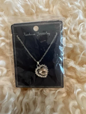 Halsband med hjärthänge och stenar - Snyggt halsband i silverfärg med ett hjärtformat hänge dekorerat med gnistrande stenar runt om. Kedjan är tunn och elegant, och hänget ger en romantisk vibe. Perfekt accessoar för dig som gillar detaljer och vill ha något som sticker ut.