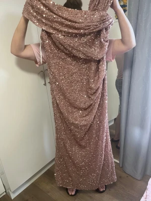 Glittrig långklänning i rosa - Säljer en superfin långklänning i rosa med massor av glittriga paljetter. Klänningen har en elegant drapering och är perfekt för dig som vill sticka ut. Materialet känns mjukt och följsamt, och den har en klassisk, rak passform. 