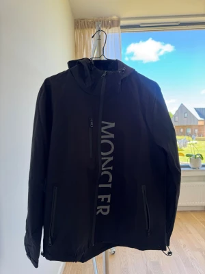 Svart Moncler vindjacka med huva - Snygg svart vindjacka från Moncler med stor logga längs dragkedjan framtill. Jackan har huva, tre dragkedjeförsedda fickor och justerbar nederkant. Perfekt för blåsiga dagar och stilren streetwear-look.