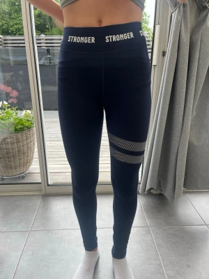 Mörkblå leggings från Stronger - Snygga mörkblå leggings från Stronger med hög midja och logga i vitt runt linningen. De har en vitprickig detalj runt ena låret som ger en sportig look. Tillverkade i stretchigt funktionsmaterial som sitter skönt och formar sig efter kroppen. Man kan vika ner midjan för att ha en snygg låg midjad look.
