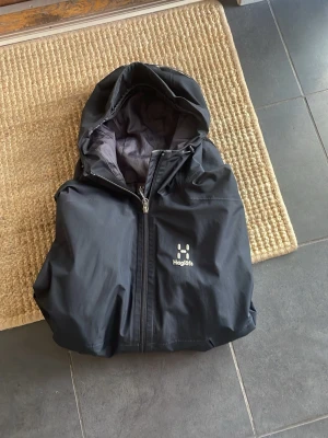Svart regnjacka från Haglöfs - Säljer en svart regnjacka med en justerbar huva. Lite dyrare exemplar då nypris på denna jacka ligger på 3799kr och jackan är knappast använd och i toppform. Skicka gärna prisförslag är öppen för förhandling!