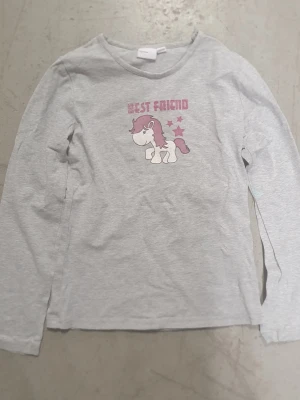 Tröja - Ljusgrå långärmad tröja i mjuk bomull med tryck av en gullig enhörning och texten 'Best Friend' i rosa på bröstet. Tröjan har rund halsringning och avslappnad passform. Perfekt för dig som gillar söta motiv och enkel stil. Finns små fläckar.