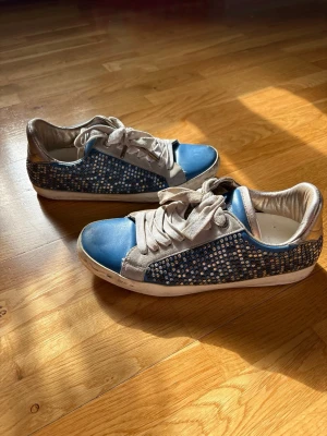 Blå sneakers med strassdetaljer från Zadig & Voltaire  - Säljer ett par blå sneakers med glittriga strassdetaljer på sidorna och vita snören. Skorna har en låg profil och vit sula. Perfekta för dig som vill sticka ut med lite extra bling i vardagen. Materialet är en mix av textil och syntet.