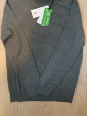 Grå stickad tröja från Lindbergh - Mjuk och stilren grå stickad tröja från Lindbergh i slim fit-modell. Tröjan har rund halsringning och ribbade muddar vid ärmslut och nederkant. Tillverkad i EcoVero-material för en mer hållbar stil. Färgen är dark moss.