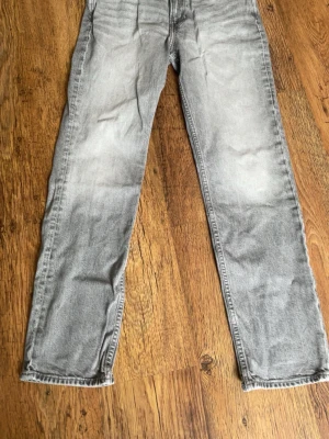 Grå tvättade raka jeans - Säljer ett par grå tvättade jeans med rak passform och klassisk femficksdesign. Jeansen har en snygg sliten look och är perfekta för en avslappnad stil. Materialet är denim i bomull och färgen är ljusgrå med tvättade detaljer.