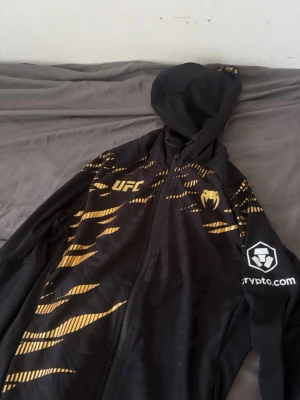  UFC hoodie med  - Snygg svart hoodie från Venum x UFC med dragkedja, huva och guldiga detaljer. Tryck med UFC-logga, Venum-logga och Crypto.com på ärmen. Tillverkad i ett lätt och sportigt material, perfekt för dig som gillar streetwear och sportig stil.