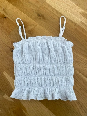 Vit broderad linnetopp med volang - Söt vit linnetopp med smala axelband och broderade detaljer. Toppen har smockad resår och volangkant nedtill för en romantisk vibe. Perfekt för varma dagar och enkel att matcha med jeans eller kjol.