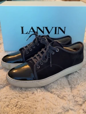 Lanvin svarta sneakers med lackdetalj - Snygga svarta sneakers från Lanvin med klassisk låg silhuett. Skorna har ovandel i mocka och en glansig lackad tå, samt vita gummisulor. Matchande svarta skosnören och metallfärgade öljetter ger en stilren look.