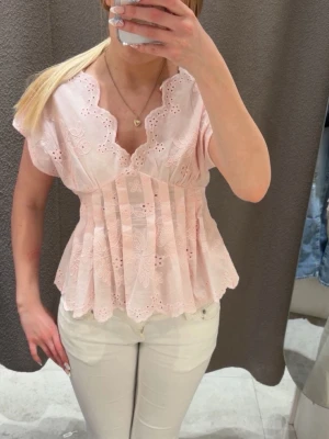 Ljusrosa broderad blus med volang - Supersöt ljusrosa blus med broderade blommor, v-ringning och volangkant. Blusen har korta ärmar och en feminin, lätt utsvängd passform. Perfekt för dig som gillar romantisk och söt stil.