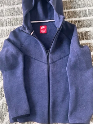 Mörkblå zip hoodie från Nike - Snygg mörkblå hoodie från Nike med dragkedja framtill och huva. Klassisk design med diskret Nike-logga på insidan och mjukt material som känns skönt mot huden. Perfekt för chill dagar eller när du vill ha en sportig look.