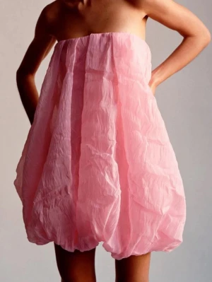 Rosa axelbandslös ballongklänning - Unik rosa axelbandslös kortklänning med ballongformad kjol och skrynkligt tyg. Perfekt för dig som vill sticka ut med en lekfull och trendig look. Klänningen har en luftig passform och är superenkel att styla. Använd en gång