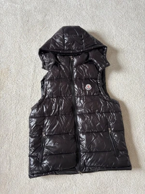 Svart dunväst från Moncler med huva - Snygg svart dunväst från Moncler med glansig finish, huva och två dragkedjefickor. Västen har Moncler-logga på bröstet och quiltad design som ger en modern look. Perfekt för lager-på-lager och streetstyle.