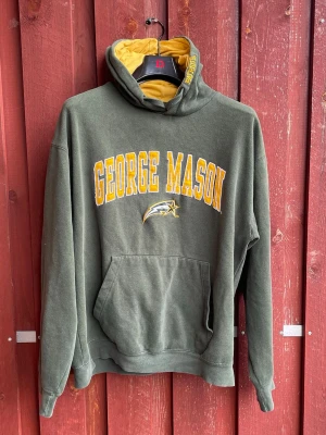 George Mason grön hoodie - Mörkgrön hoodie med gul insida i huvan och stor gul text 'GEORGE MASON' på bröstet. Klassisk känguruficka framtill och tryckt logga under texten. Mjuk bomullskänsla och avslappnad passform, perfekt för college-vibe. Söm har spruckit något i ena armhålan
