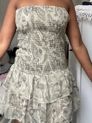 Beige axelbandslös kortklänning - Säljer en beige axelbandslös kortklänning med volangkjol och smockad överdel. Klänningen har ett paisleyinspirerat mönster och är perfekt för varma dagar. Materialet känns lätt och luftigt, vilket gör den skön att bära.