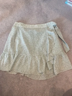 Grön omlottkjol med volang - Söt grön omlottkjol med vitt blommönster och volangkant. Kjolen har knytband i midjan och är tillverkad i ett lätt och luftigt material, perfekt för varmare dagar. Passar dig som gillar en feminin och trendig stil.