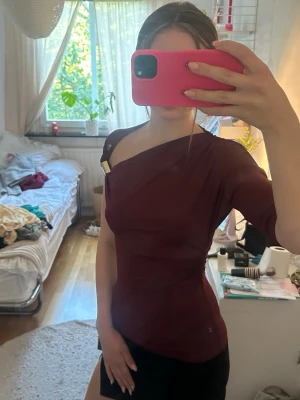 Vinröd oneshoulder topp med detalj - Snygg vinröd topp med oneshoulder-design och guldfärgad metallspänne på axeln. Tight passform som framhäver figuren och kort ärm på ena sidan. Perfekt för dig som vill sticka ut med en unik och trendig look.