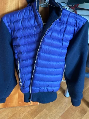 Lila cardigan från LeVon - Snygg lila cardigan från LeVon med quiltad front och rygg i blankt material samt svarta ärmar och muddar i mjuk fleece. Jackan har dragkedja framtill och en sportig look som passar perfekt till kalla vinterdagar. En knapp har låsnat vid luvan. Pris kan såklart diskuteras!