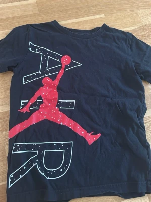 Svart Air Jordan t-shirt - Snygg svart t-shirt från Jordan med stort AIR-tryck och den klassiska Jumpman-loggan i rött framtill. T-shirten har rund hals och korta ärmar. Perfekt för dig som gillar streetwear och sportig stil.
