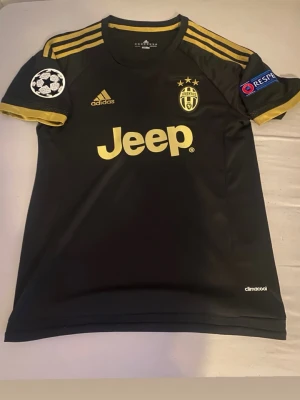 Juventus Dybala fotbollströja - Juventus fotbollströja i svart med guldiga detaljer, Adidas-logga och Jeep som sponsor. Tröjan har Dybala och nummer 21 på ryggen, Champions League-märken på ärmarna och tre stjärnor ovanför klubbmärket. Tillverkad i lätt och ventilerande climacool-material.