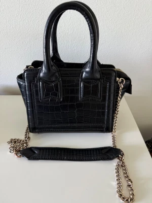 Svart handväska med kedjerem - Snygg svart handväska i croco-mönstrat konstläder med guldfärgad kedjerem och dubbla handtag. Väskan har en fyrkantig form och dragkedja upptill. Perfekt accessoar för dig som gillar trendiga detaljer och stilren design.