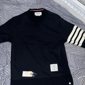 Thom Browne Sweater  - Svart långärmad topp från Thom Browne med vita ränder på ena ärmen och en vit patch med märkets logga framtill. Rund hals och diskret röd, vit och blå detalj i nacken. Tillverkad i mjuk bomull för en clean och stilren look. Storlek S men lite för liten, passar mera XS. Plastpåse och Dustbag medföljer