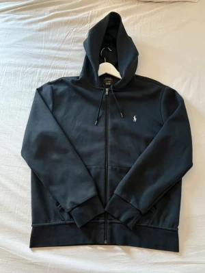 Svart hoodie från Polo Ralph Lauren - Säljer en svart hoodie från Polo Ralph Lauren med dragkedja framtill och klassisk vit logga på bröstet. Hoodien har justerbar huva med snören, ribbade muddar och fickor på sidorna. Perfekt för en chill och stilren look.