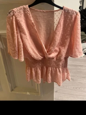 Ljusrosa blus med v-ringning och volang - Supersöt ljusrosa blus med v-ringning, puffiga korta ärmar och volang nedtill. Tyget är lätt och har små prickiga detaljer för extra textur. Smockad midja ger en snygg passform och toppen känns både luftig och trendig.