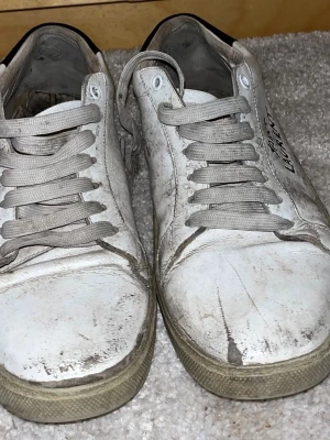 Vita Saint Laurent sneakers - Klassiska vita sneakers från Saint Laurent med handskriven logga på sidan. Skorna har låga skaft, vita snören och ovandel i läder. Snygg minimalistisk design som passar till det mesta. Perfekta för dig som gillar exklusiva märken och streetstyle.