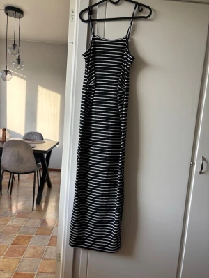 Svartvit långrandig klänning - Långklänning med smala axelband i svart och vitt randigt mönster. Klänningen har en rak passform och är tillverkad i ett mjukt, stretchigt material som känns bekvämt mot huden. Perfekt för varma dagar och enkel att styla.