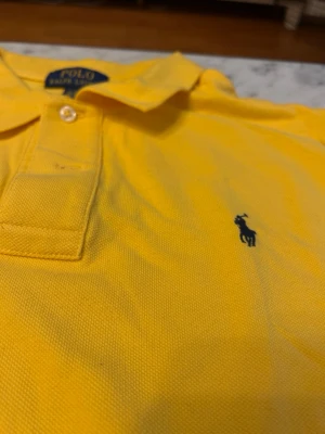 Gul piké Polo Ralph Lauren  - Klassisk gul pikétröja från Polo Ralph Lauren i storlek XL. Tröjan har krage, knappar framtill och den ikoniska broderade loggan på bröstet. Tillverkad i mjuk bomull som känns skön mot huden. Perfekt för en avslappnad och stilren look.
