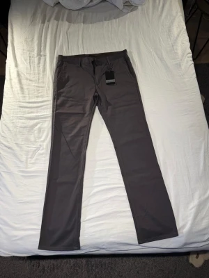Mörkgrå chinos med raka ben - Snygga mörkgrå chinos med raka ben och klassisk passform. Byxorna har framfickor, bälteshällor och två bakfickor med lock. Tillverkade i ett mjukt och slitstarkt bomullsmaterial som ger en clean look. Perfekta för dig som gillar stilrena och enkla byxor.