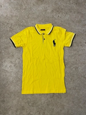 Gul piké från Ralph Lauren - Klassisk gul pikétröja från Ralph Lauren med svart logga på bröstet. Tröjan har korta ärmar, krage med svarta detaljer och knappar framtill. Tillverkad i mjuk bomull som känns skön mot huden. Perfekt för en avslappnad och stilren look.