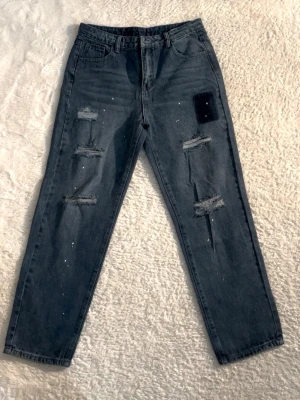 Svarta slitna jeans med färgstänk, finns i shorts! - Säljer ett par svarta jeans med raka ben, slitningar och hål på både fram- och baksida samt vita färgstänk för en edgy look. Jeansen har klassisk femficksmodell och hög midja. Perfekta för dig som gillar streetstyle och vill sticka ut.