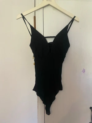 Svart ribbad body med smala axelband - Svart body från Vero Moda med ribbad struktur och smala axelband. Djup v-ringning framtill och dekorativ rem över bröstet. Stretchigt material som sitter tight mot kroppen. Perfekt för att styla med jeans eller kjol.